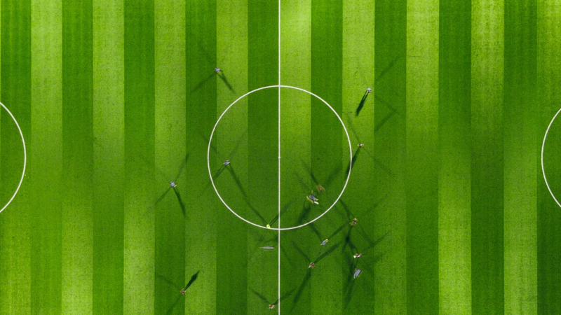 The image shows a football field from above. The players are only visible because of their shadows, symbolizing Humans in the Loop.