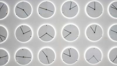 The picture shows a white wall with several clocks, all showing a different time. This symbolises the paradoxical impact of generative AI in the workplace on productivity.