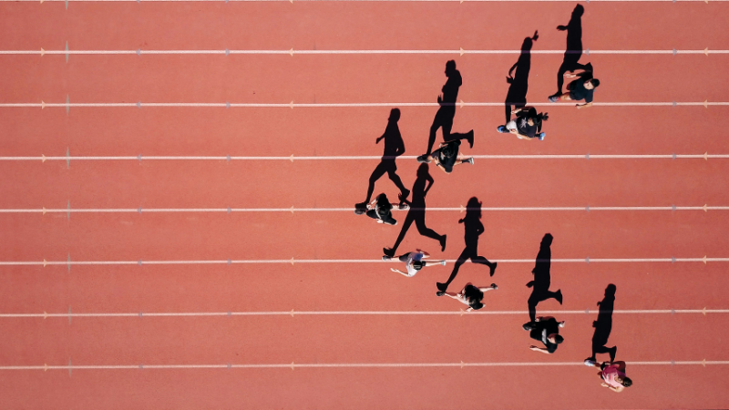 Das Foto zeigt eine Gruppe Läufer*innen von oben. Sie werfen lange Schatten auf die Rennbahn und bilden von oben eine Pfeilformation. Diese steht symbolisch dafür, dass KI zwar als Zukunftstechnologie gefeiert wird, aber derzeit vor allem die Vergangenheit vorhersagt.