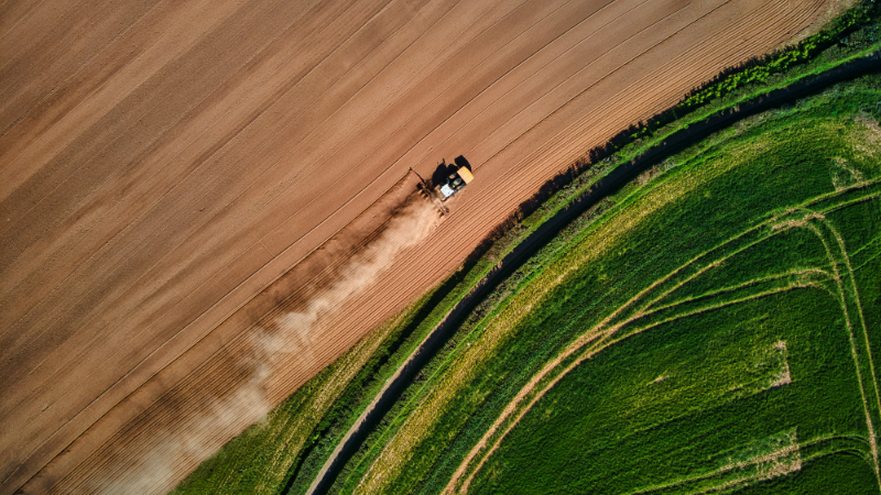 The picture shows a tractor cultivating a field from above. One side of the field is covered in green, the other is dry and earthy. This is intended to show that although sustainable AI can be useful in the fight against climate change, it also comes at a high ecological cost.