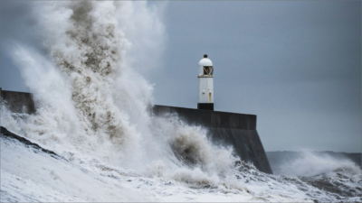 A powerful image symbolising resilience: ocean waves crashing against a lighthouse during the daytime, representing researchers standing strong against science hostility.