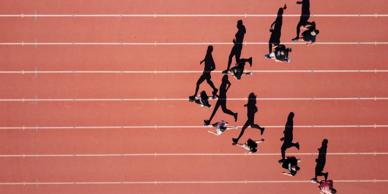 Das Foto zeigt eine Gruppe Läufer*innen von oben. Sie werfen lange Schatten auf die Rennbahn und bilden von oben eine Pfeilformation. Diese steht symbolisch dafür, dass KI zwar als Zukunftstechnologie gefeiert wird, aber derzeit vor allem die Vergangenheit vorhersagt.