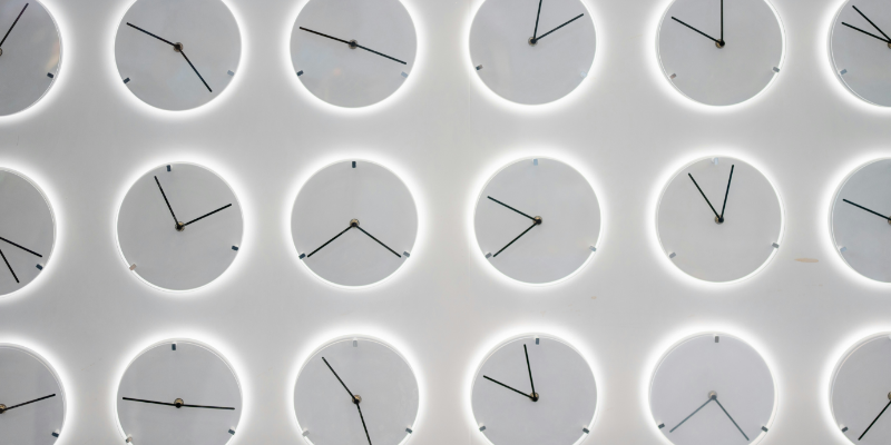 The picture shows a white wall with several clocks, all showing a different time. This symbolises the paradoxical impact of generative AI in the workplace on productivity.