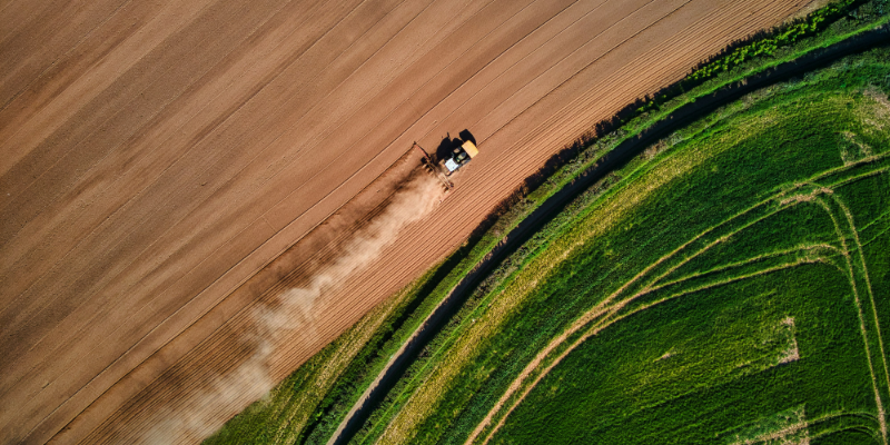 Das Bild zeigt einen Traktor von oben, der ein Feld bestellt. Eine Seite des Feldes ist grün bewachsen, die andere trocken und erdig. Das soll zeigen, dass nachhaltige KI zwar im Kampf gegen den Klimawandel nützlich sein, selbst aber auch hohe Kosten für die Umwelt verursacht.